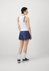 Balts tops bez piedurknēm, tumši zils svārku modelis ar platu apakšu, melni sporta apavi un balti zeķīši. Gluda auduma, plūstošs dizains un minimālistiski sīkumi.