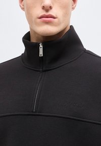 Άνδρας που φοράει μαύρο φούτερ με φερμουάρ και ψηλό γιακά, με διακριτικό λογότυπο Calvin Klein στο στήθος, πάνω σε απλό φόντο.