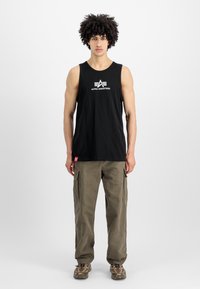 Μαύρο μπλουζάκι κοντομάνικο με λευκό λογότυπο "Alpha Industries", συνδυασμένο με ελαιόχρωμα cargo παντελόνια που διαθέτουν μεγάλες πλαϊνές τσέπες και ρεβέρ στα μπατζάκια.