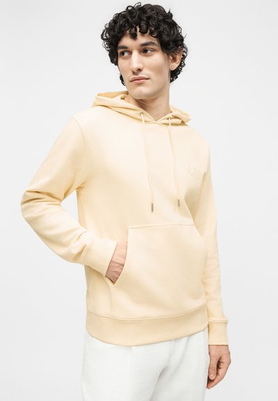 Νέος άνδρας με σγουρά μαλλιά που φοράει ανοιχτό μπεζ φούτερ με τσέπη μπροστά και λευκό παντελόνι, κοιτάζοντας προς τα πλάγια σε ένα απλό φόντο.