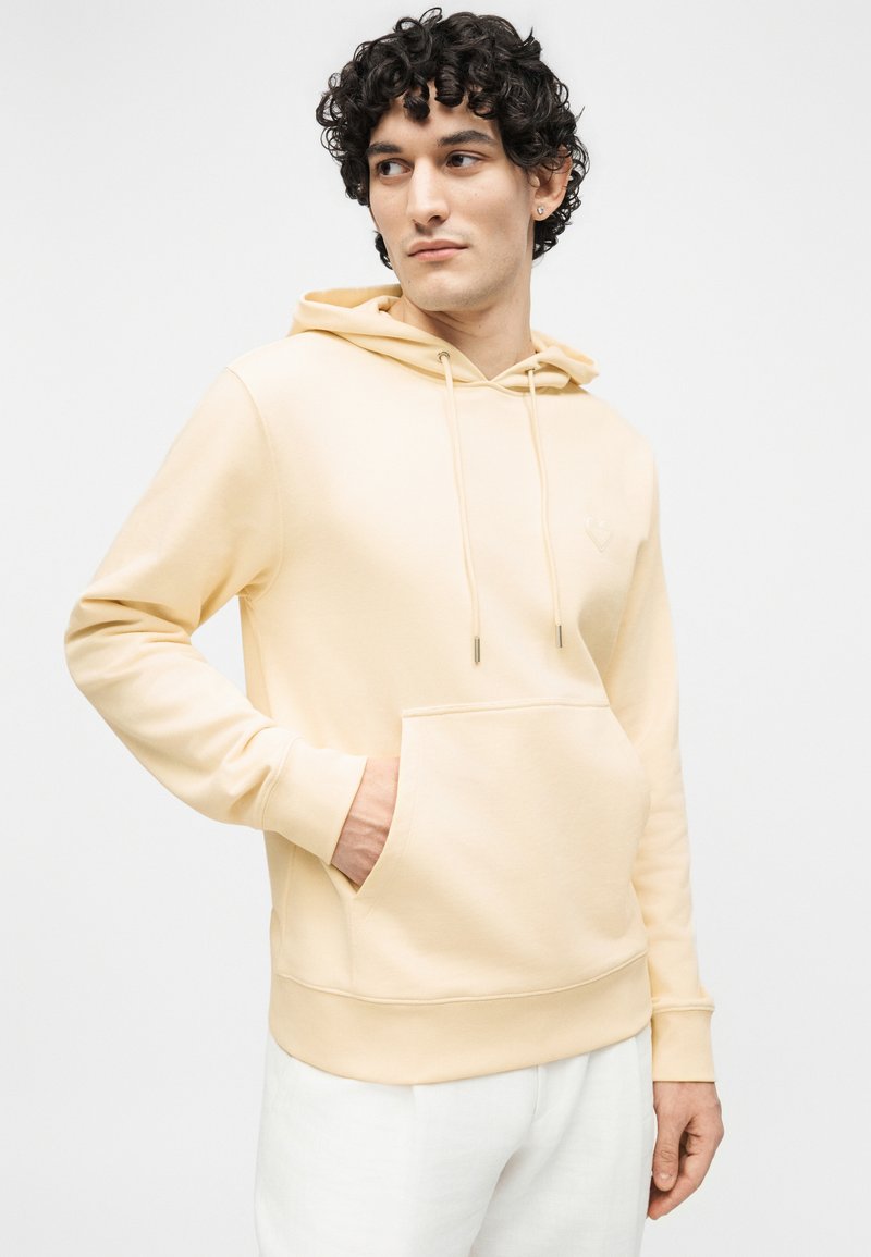 Young man with curly hair wearing a light beige hoodie with front pocket and white pants, looking to the side against a plain background.