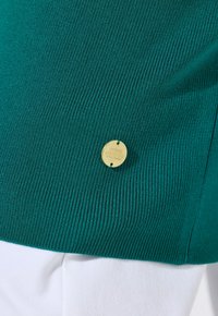 Maglione verde a coste con una finitura testurizzata, caratterizzato da un'etichetta rotonda dorata con testo in rilievo, abbinato a un tessuto bianco sottostante.