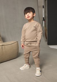 Beiges Sweatset bestehend aus einem Sweatshirt mit Rundhalsausschnitt und schmal zulaufenden Jogginghosen. Das Outfit hat gerippte Bündchen und ein kleines Logodetail. Weiße Sneaker mit Akzenten.