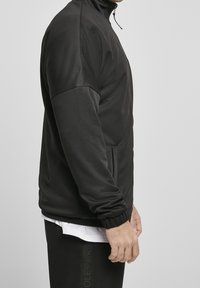 Veste de sport noire avec un col haut, des manches longues et une fermeture éclair à l'avant. Elle possède une texture lisse et des poches latérales.