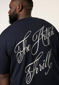 Marineblå bomuld t-shirt med stort hvidt kursiv tekst "The Artful Thrill" trykt henover ryggen. Løs pasform og korte ærmer.