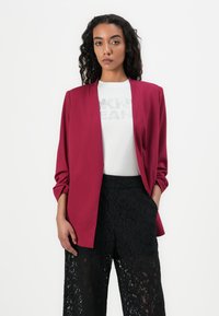 Blazer bordeaux avec une texture lisse, devant ouvert, et manches longues, associé à un T-shirt graphique blanc et un pantalon large en dentelle noir.