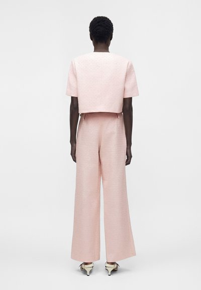 Personne aux cheveux courts et bouclés portant un haut rose clair à manches courtes et un pantalon ample, debout, dos tourné sur un fond blanc.