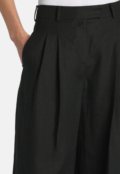 Kéz egy magas derekú, pliszírozott fekete bő szárú nadrág zsebében, fehér háttér előtt.