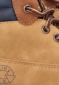 Botas de ante tan con acentos marrones y tejido azul marino. Presenta ojales de metal, cordones trenzados y un logo circular en el lateral.