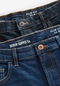 Slim fit traperice u tamnoplavoj boji. Imaju mesingane dijelove, kontrastne šavove i oznake s "DENIM SUPPLY Co." i informacijama o veličini.