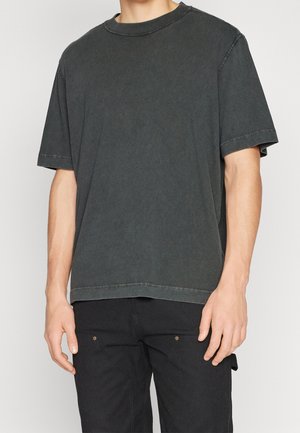 Mann trägt ein verblasstes schwarzes, oversized T-Shirt mit kurzen Ärmeln und schwarze Hose, steht vor einem schlichten hellen Hintergrund.