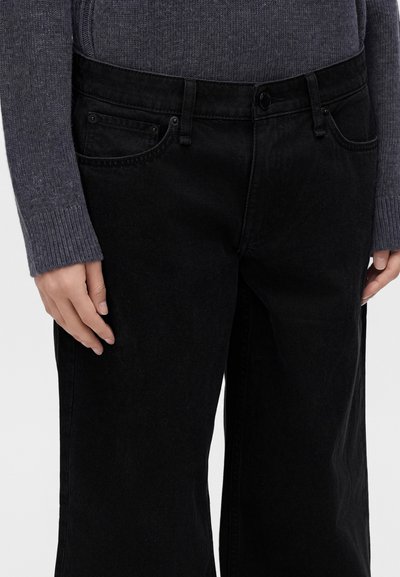Personne portant un jean noir à jambes larges et un pull côtelé gris foncé, les mains reposant sur les côtés, vue recadrée de la partie médiane.