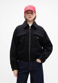 Sort fleecejakke med lynlås foran og stor krave, med to brystlommer. Sammen med mørke denimbukser og en rød kasket.