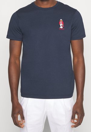 Mann trägt ein marineblaues T-Shirt mit einem kleinen bestickten Bären in rot-weißem Outfit auf der linken Brust, kombiniert mit weißen Hosen.