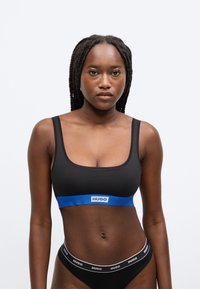 Musta urheiluliivit, joissa on leveä sininen joustava nauha, jossa on "HUGO"-logo. Kangas näyttää sileältä ja joustavalta, ja siihen on yhdistetty mustat alushousut.