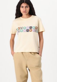 Crèmekleurig katoenen t-shirt met een ruitpatroon van bloemmotieven in blauw, groen, paars en bruin over de borst. Korte mouwen, comfortabele pasvorm.