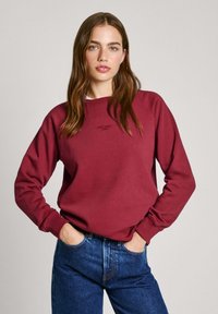 Felpa borgogna con collo a giro e polsini a coste, abbinata a jeans in denim blu. Presenta un piccolo logo al centro.