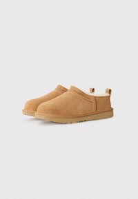 Scarpe slip-on in suede tan con morbida fodera in pelliccia, profilo basso, suola testurizzata e dettagli cuciti lungo il bordo. Design leggero e informale.