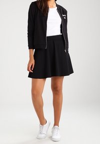 Vrouw gekleed in een zwarte zip-up jas, wit shirt, zwarte rok en witte sneakers, staand tegen een effen achtergrond.