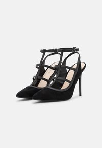 Sapatos de salto alto em camurça preta, com um design aberto atrás, bicos apontados e várias tiras cruzadas. Apresentam uma fivela no tornozelo para ajuste.