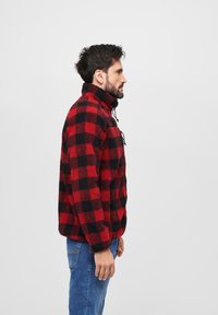 Rød og sort ternet fleece-pullover med høj krave, lynlås lukning og to forlommer, båret med lyseblå jeans.