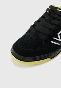 Melns zamšs sneakers ar melnām auklām, baltu zīmolu un teksturētu dzeltenu gumijas zoli. Iezīmē zemu profilu un mīkstu apkakli.
