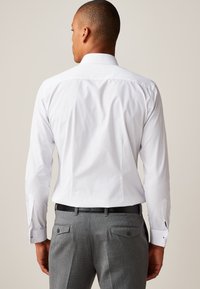 Hvid skjorte med lange ærmer, glat tekstur og en spids krave. Kombineret med grå bukser med et diskret mønster og baglommer.