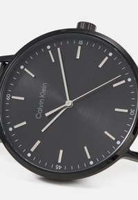 Calvin Klein svart klokke med matt urskive, sølvfargede timeindekser og enkle visere. Inkluderer en teksturert overflate og en krone på siden.