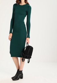 Femme portant une robe midi à manches longues côtelées vertes, tenant un petit sac à dos noir texturé et portant des bottines noires.