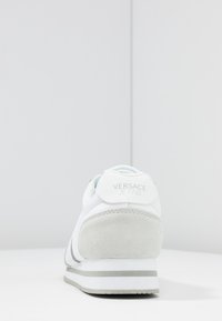 Baskets blanches avec des accents en daim gris, matériau texturé, talon arrondi et logo à l'arrière. Comprend une semelle intermédiaire rayée et un devant à lacets.