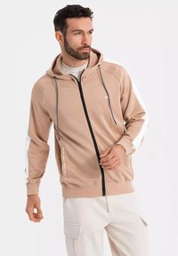 Felpa beige con zip e cappuccio regolabile, zip nera e strisce laterali bianche. Tessuto leggero, con dettagli del logo sulle maniche.