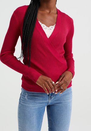 Vrouw die een geribbelde rode wikkeltrui draagt over een witte kanten top en lichtblauwe spijkerbroek, met gevlochten haar en ringen aan haar vingers.