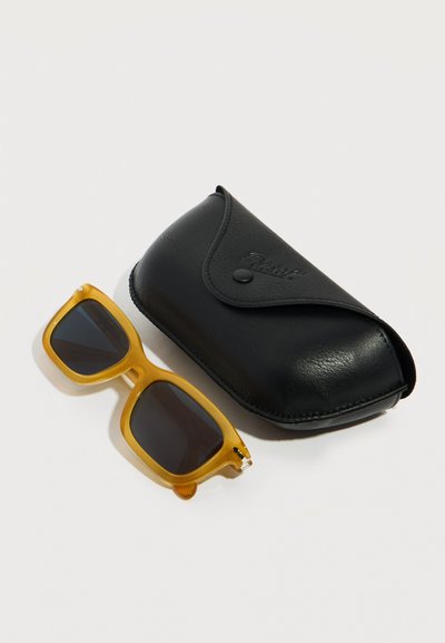 Κίτρινου πλαισίου ορθογώνια γυαλιά ηλίου με σκοτεινούς φακούς δίπλα σε κλειστή μαύρη δερμάτινη θήκη γυαλιών ηλίου πάνω σε λευκή επιφάνεια.