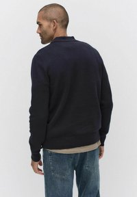 Marineblauwe trui met een kraagontwerp, ribgebreide mouwen en zoom. Draagt in combinatie met lichtdenim jeans, die een soepele textuur en een relaxte pasvorm tonen.