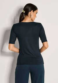 Vrouw met donker haar in een paardenstaart, die zilveren oorbellen draagt, een strakke donkerkleurige top met korte mouwen en bijpassende donkere broek, met de rug naar ons toegekeerd.