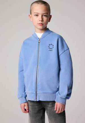 Light blue zip-up hoodie made of soft cotton with ribbed cuffs, featuring a logo print in black on the left chest. Styled with gray jeans.