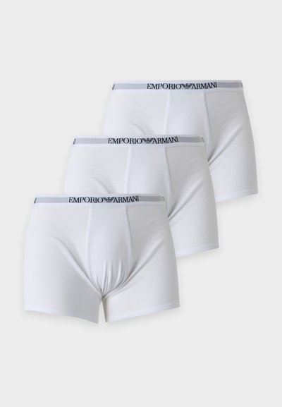 Τρία άσπρα ανδρικά boxer με γκρι ζώνες Emporio Armani διατεταγμένα διαγώνια σε ένα φωτεινό φόντο.