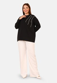 Felpa nera con dettagli in perle in un motivo ondulato, abbinata a pantaloni bianchi a gamba larga e scarpe con punta affilata. Tessuto dalla texture liscia.