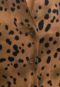 Chemise marron avec un motif léopard composé de taches noires et des fermetures à boutons. Le tissu semble lisse et léger.