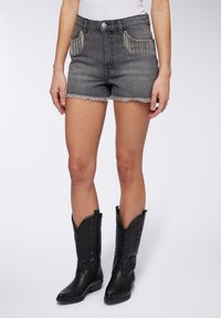 Shorts in denim grigio con orlo sfilacciato e tasche decorative con perline, abbinati a stivali da cowboy in pelle nera con dettagli in rilievo.