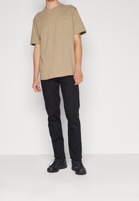 Beige bomulls t-shirt med kort ärm och bröstficka, tillsammans med svarta raka byxor och svarta träningsskor.