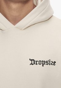 Kreemipesu, millel on eesmine kängurusoe tasku, ja millel on rinnal musta tikandkirja "Dropsize" gotikastiilis.