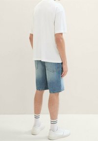 Mies, joka pukeutuu valkoiseen T-paitaan, farkkuhousuihin, valkoisiin sukkiin, joissa on mustia raitoja, ja valkoisiin lenkkareihin, seisoo yksinkertaista vaaleaa taustaa vasten.