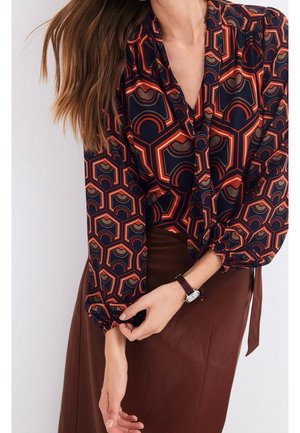 Femme portant un chemisier à motif géométrique avec des nuances d’orange, de marine et de marron, associé à une jupe marron et une montre-bracelet.