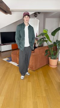 Personne portant une chemise verte à boutons sur un t-shirt blanc, un pantalon noir ample et des chaussures argentées, se tenant dans un salon moderne.