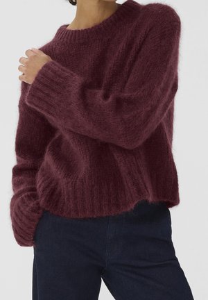 Person trägt einen flauschigen, bordeauxroten Pullover mit langen Ärmeln und dunkle Hose, mit einer Hand die gegenüberliegende Schulter berührend und der anderen in der Tasche.