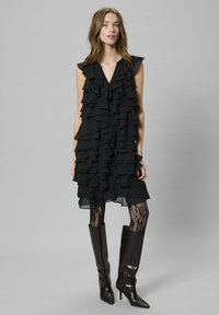 Fekete ujjatlan ruha réteges fodrokkal, áttetsző szövet részletekkel. Fekete csipke harisnyával és térdig érő, hegyes orrú csizmával viselve.