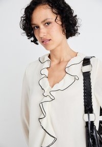 White blouse featuring a ruffled neckline with black trim. Model carries a textured black shoulder bag with a woven strap.