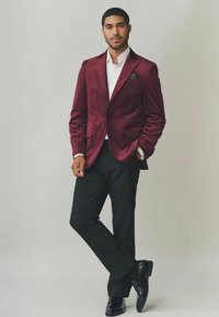 Burgunderroter Samtblazer mit Revers, schwarze Hose und glänzende schwarze Schuhe. Weißes Hemd mit einer Akzent-Taschentuch. Lässige Pose.