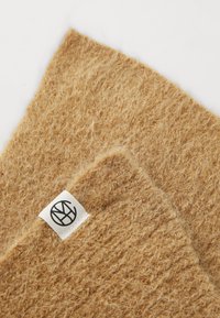 Tan wool scarf with a textured surface, featuring a woven pattern and a small white label with a black logo on one edge.